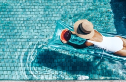 Frau in weißem Badeanzug liegt auf Luftmatratze in Hotelpool und isst Wassermelone