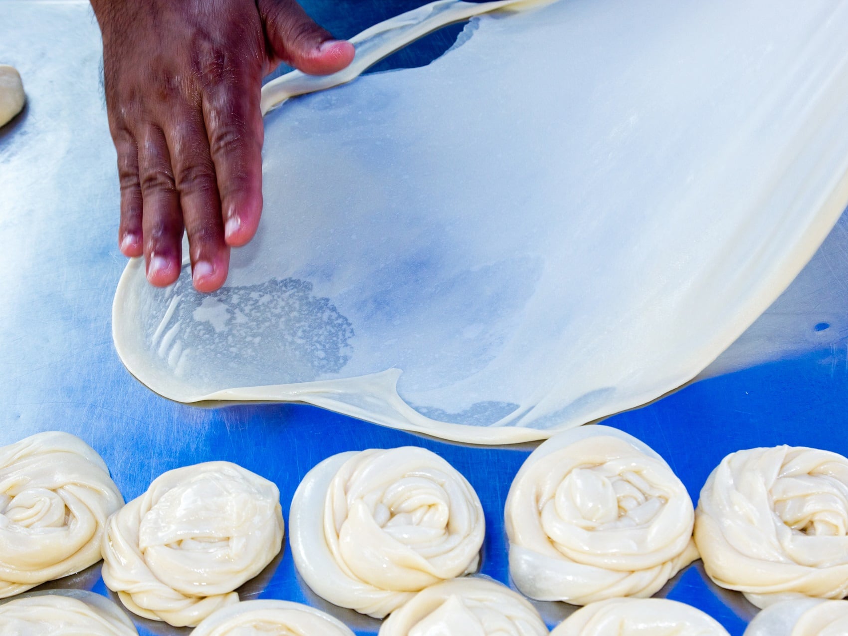 Eine Person breitet feuchten Roti-Teich zu einem großen Fladen aus. Weiter unten auf der Arbeitsfläche liegen bereits mehrere Roti zusammengerollt, bereit zum Backen.