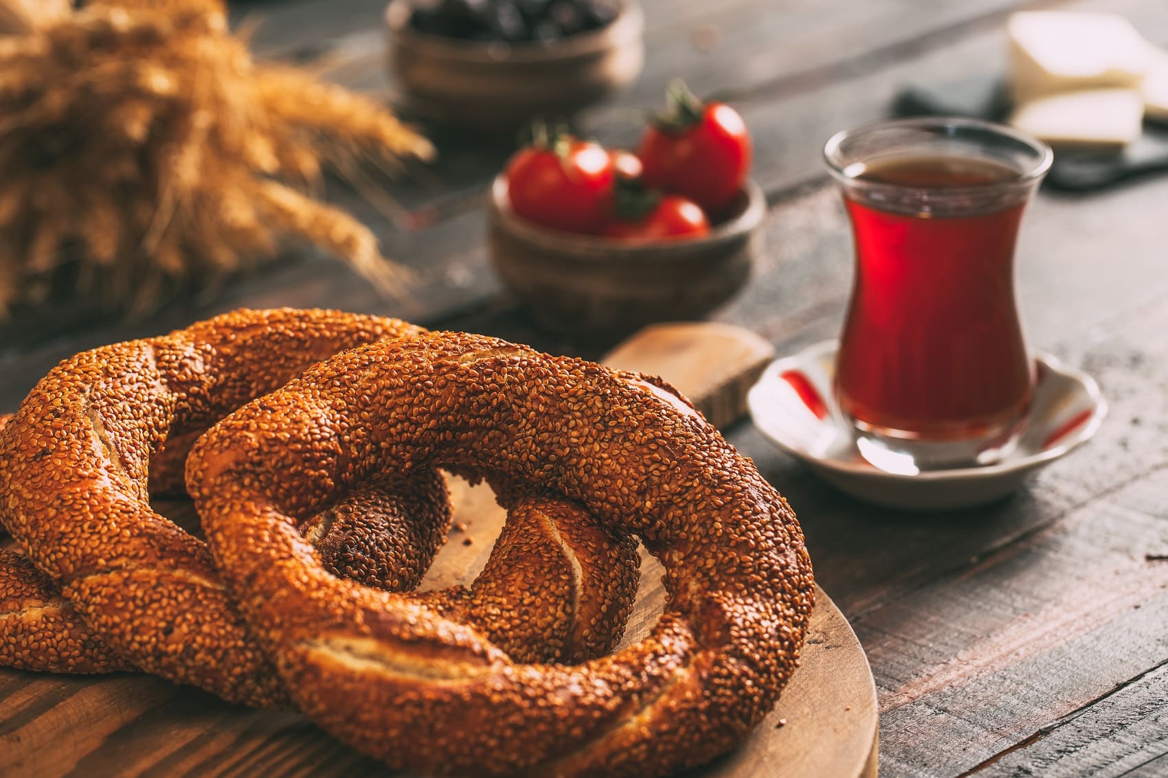 Auf einem Brettchen liegen drei Simit-Kringel, dahinter auf einem Tisch eine Tee in einem typisch türkischen Glas.