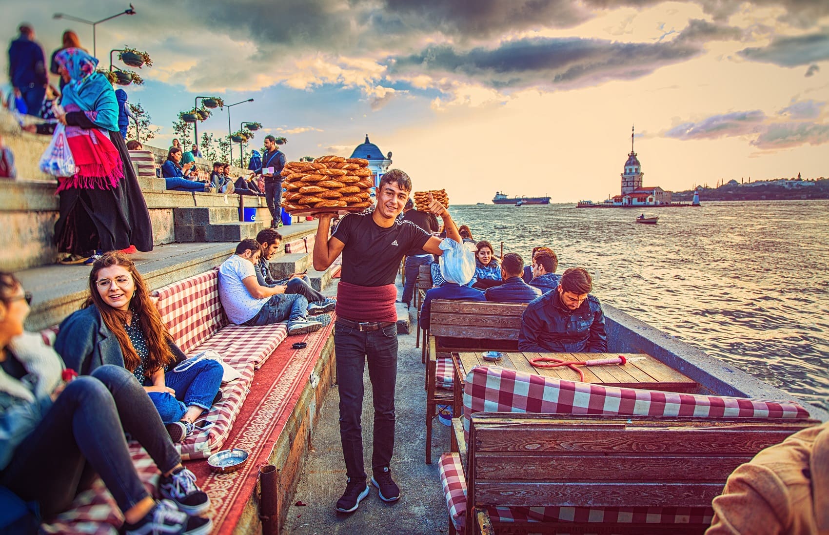 Ein Mann verkauft auf einer Promenade in Istanbul Simit; in der rechten Hand hält er ein rundes Tablett in Schulterhöhe, auf dem er gekonnt mehrere Lagen Simits balanciert.