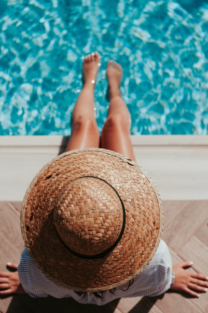 Frau mit Sonnenhut sitzt am Pool und hält ihre Füße ins Wasser