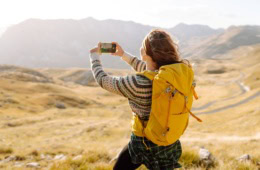 Eine junge Frau mit Outdoor-Klamotten und gelbem Rucksack macht von der vor ihr liegenden Hügellandschaft ein Foto mit ihrem Smartphone