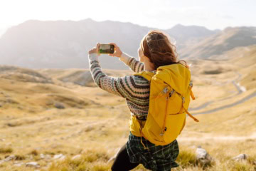 Eine junge Frau mit Outdoor-Klamotten und gelbem Rucksack macht von der vor ihr liegenden Hügellandschaft ein Foto mit ihrem Smartphone
