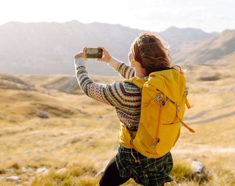 Eine junge Frau mit Outdoor-Klamotten und gelbem Rucksack macht von der vor ihr liegenden Hügellandschaft ein Foto mit ihrem Smartphone