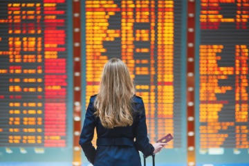 Elegant gekleidete Frau steht am Flughafen vor einer riesigen Anzeigetafel