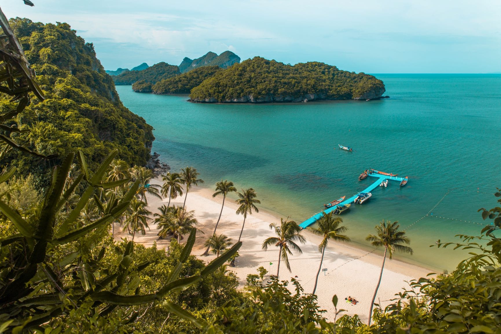 Strand im Ang Thong Marine National Park: der kleine Strandabschnitt ist von dichtem Wald umgeben, vom Ufer führt ein blauer, schmaler Steg ins Wasser, an dem einige Boote liegen.