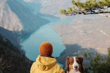 Frau in gelber Jacke und oranger Mütze sitzt mit dem Rücken zur Kamera auf einem Felsvorsprung und hält den Arm um einen Australian Shepherd, unter ihnen der Flusslauf der Drina zwischen bewaldeten Bergen.