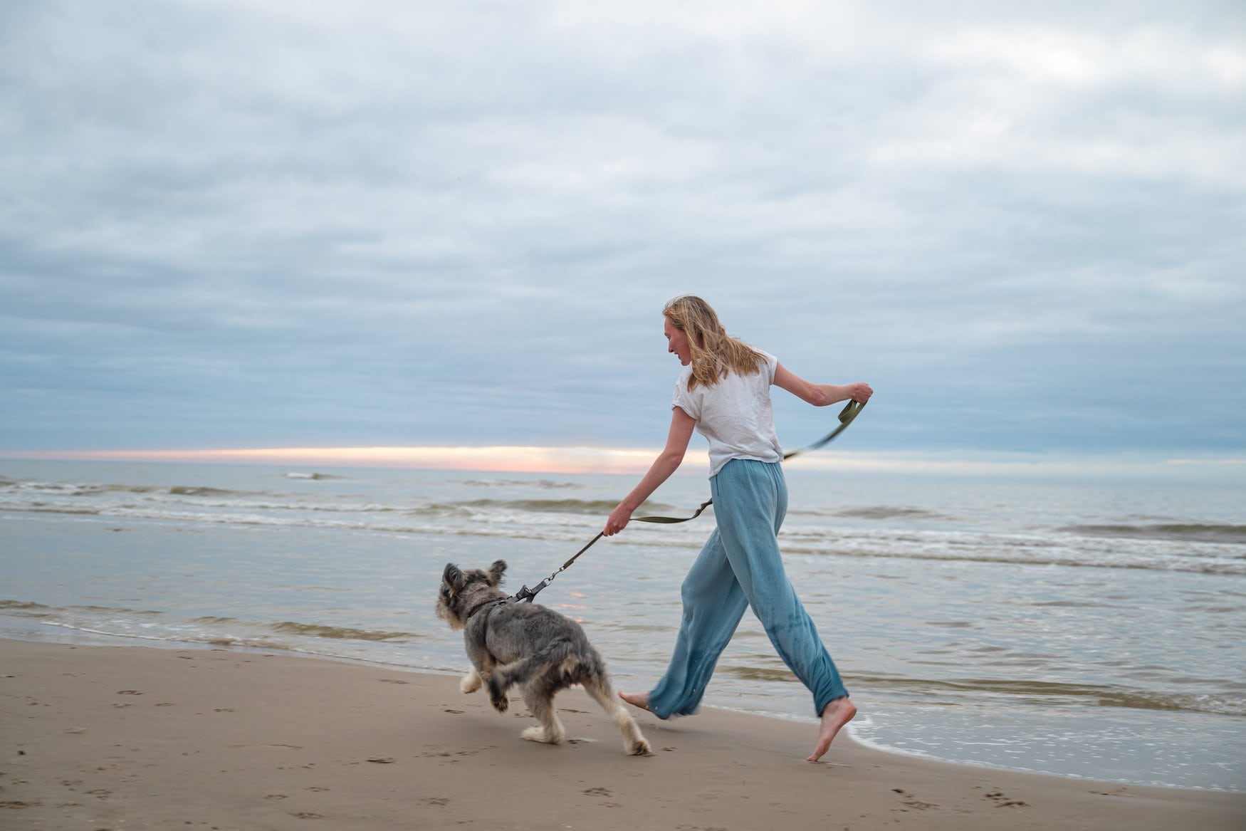 Frau in weißem Shirt und blauer Hose läuft barfuß mit einem Hund an der Leine über den nassen Sandstrand, im Hintergrund die Brandung bei bedecktem Himmel.