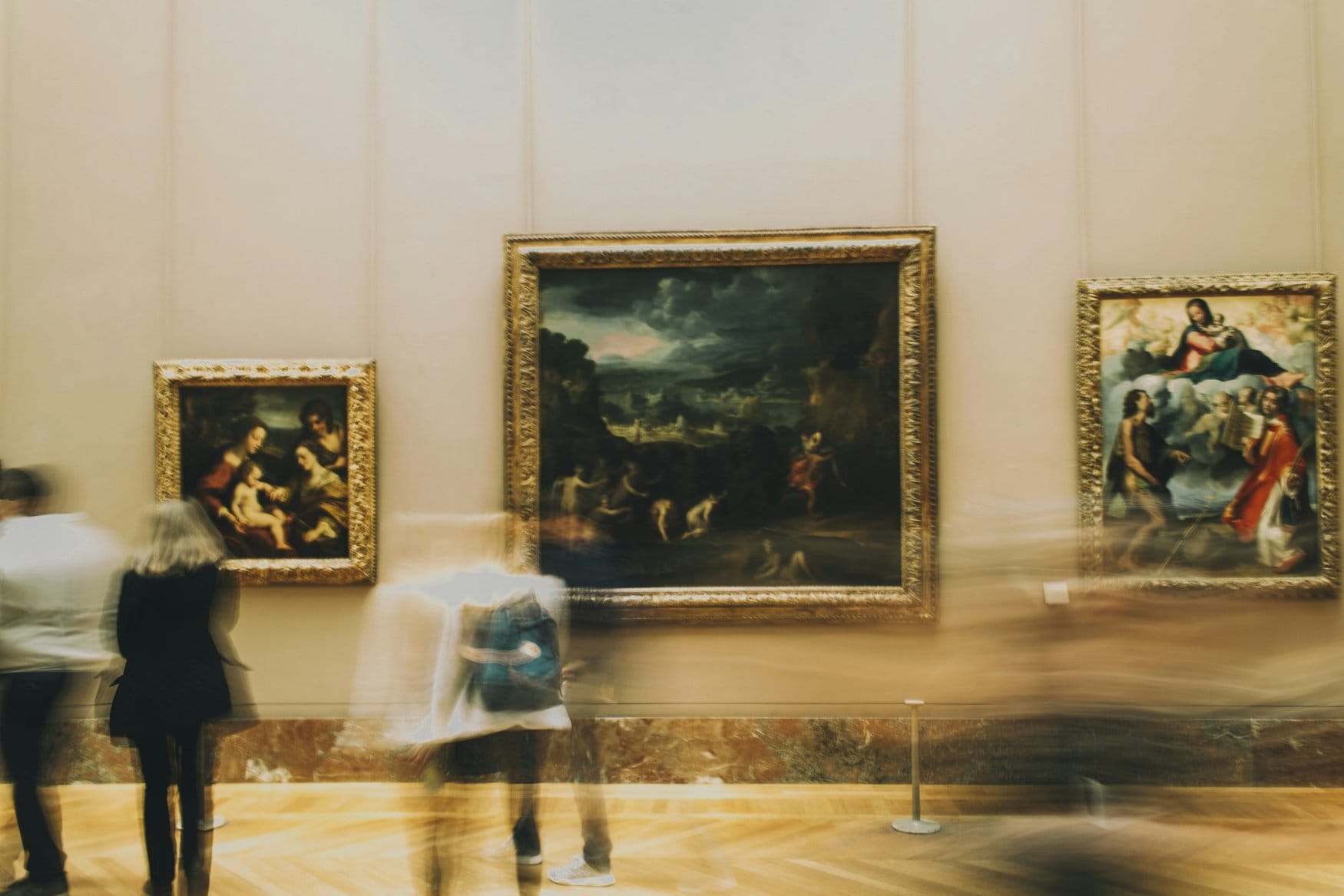 Gemäldesaal im Louvre mit drei goldgerahmten Werken an der Wand, die vorbeigehenden Besucher erscheinen durch die lange Belichtungszeit verwischt.