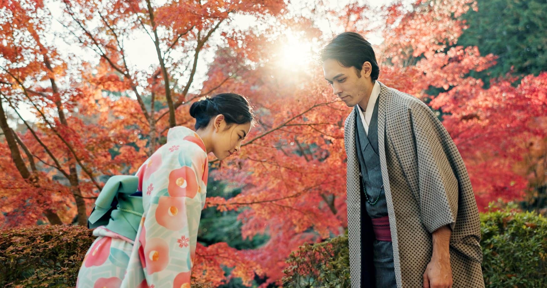 Eine Frau im Kimono verbeugt sich vor einem Mann in traditioneller japanischer Kleidung, umgeben von rot gefärbtem Ahornlaub im Gegenlicht.