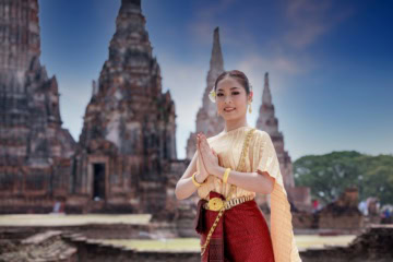 Eine Frau in traditioneller thailändischer Tracht mit goldenem Schmuck und einer Frangipaniblüte im Haar legt die Handflächen zum Wai zusammen. Hinter ihr erheben sich die Ziegeltürme des Wat Chaiwatthanaram in Ayutthaya.