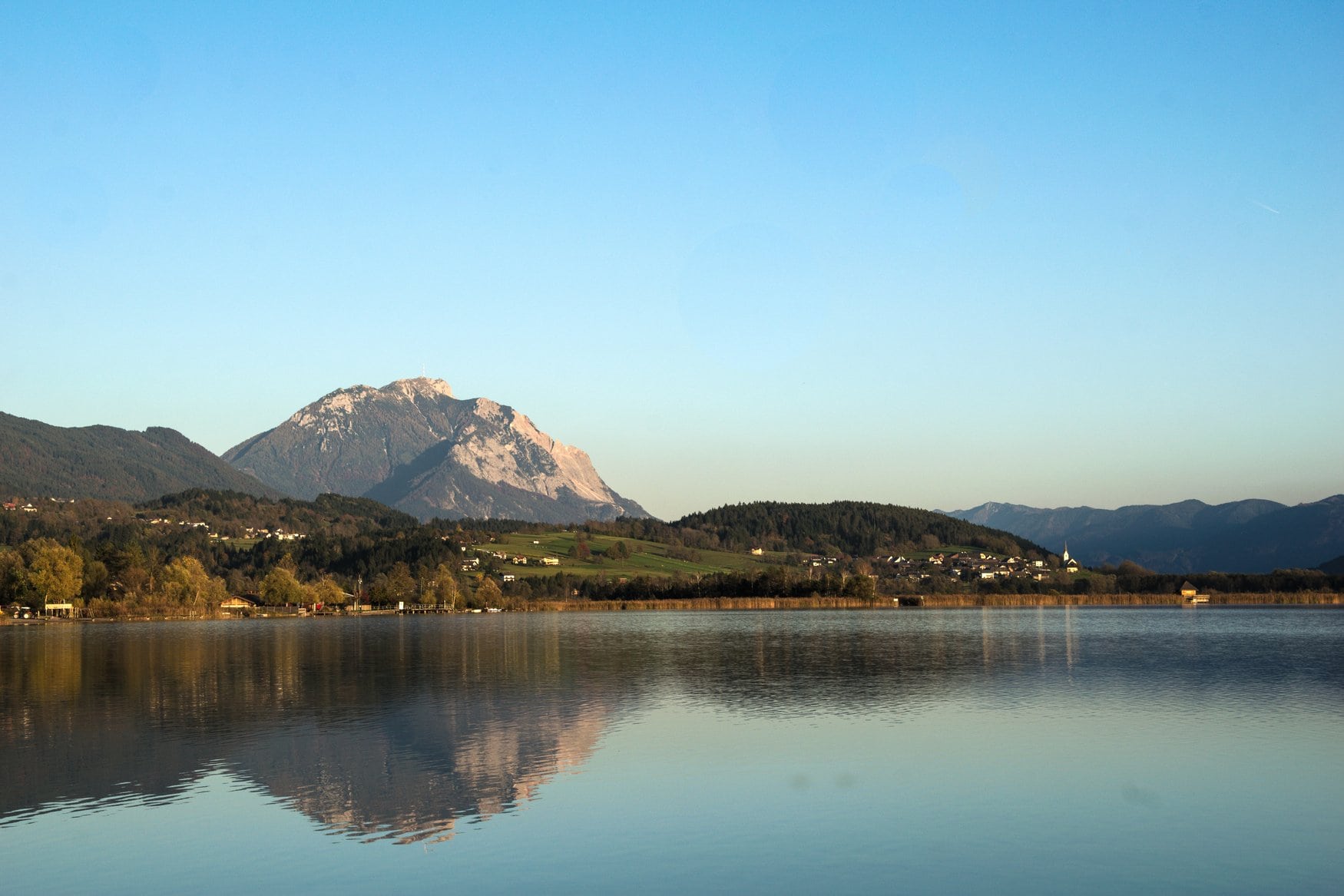 Der Pressegger See in Österreich bei ruhigem Wasser mit Spiegelung der umliegenden Berge, am Ufer ein Ort mit Kirchturm vor bewaldeten Hügeln.