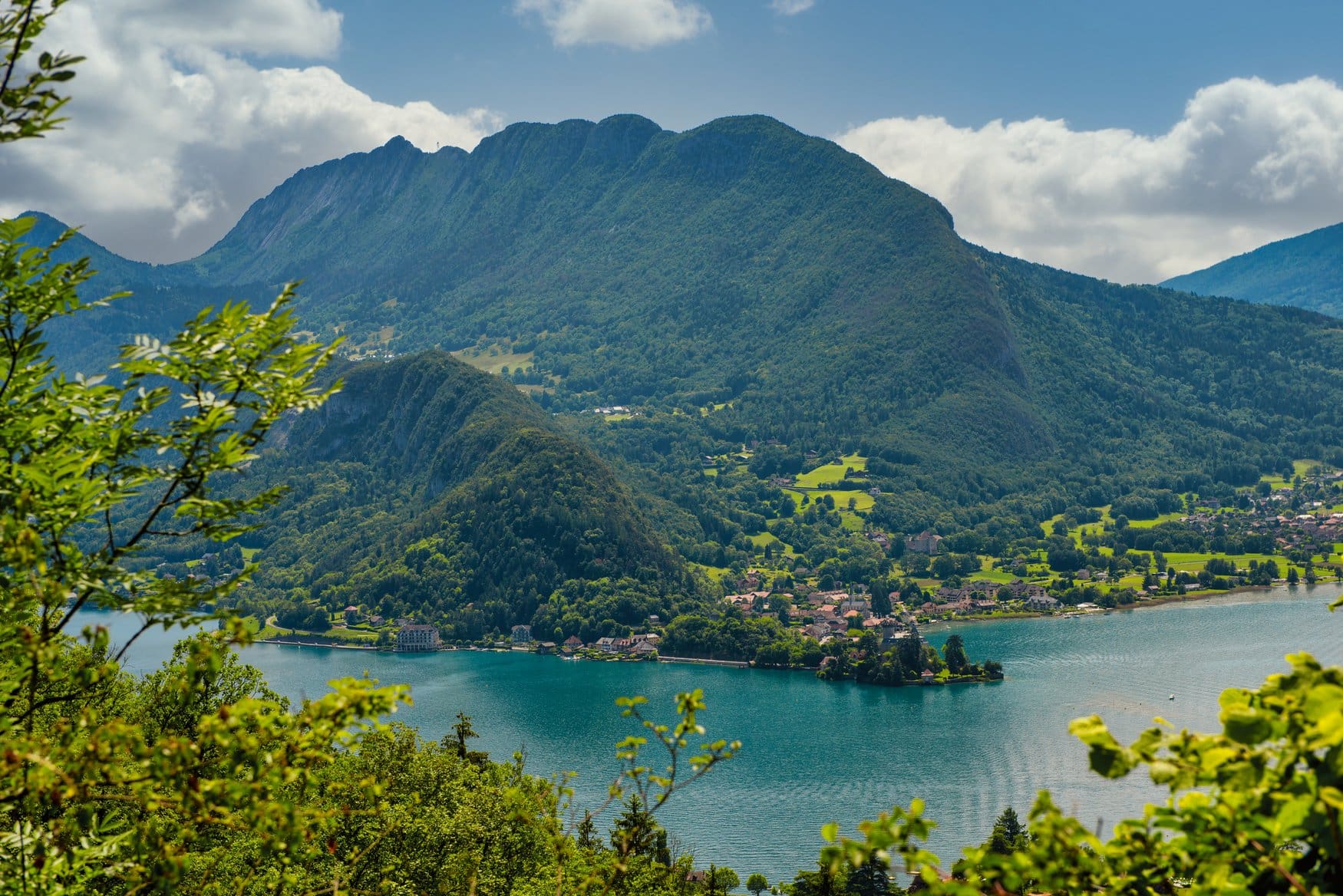 Blick über den Lac d'Annecy in Frankreich auf ein Dorf am Ufer, dahinter bewaldete Hänge und ein langgezogener Bergrücken unter teils bewölktem Himmel.