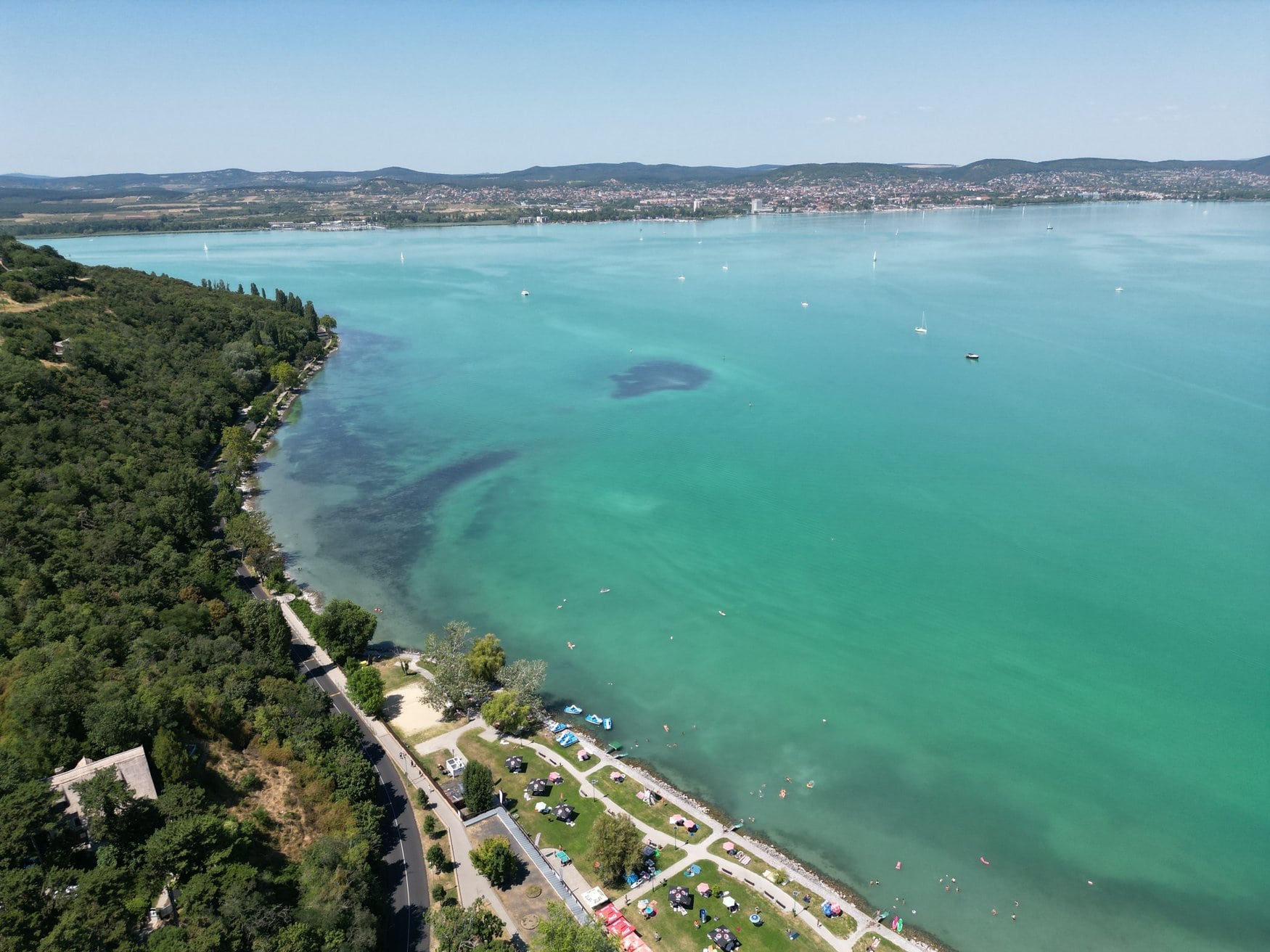 Luftaufnahme des Plattensees in Ungarn mit türkisfarbenem Wasser, einem bewaldeten Uferstreifen mit Liegewiese und einigen Segelbooten auf dem See.