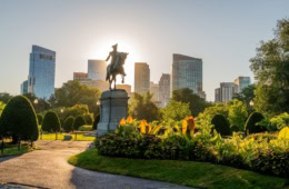 Reiterstatue im Boston Public Garden vor der Skyline der Innenstadt im warmen Abendlicht, umgeben von gepflegten Blumenbeeten und Parkwegen.