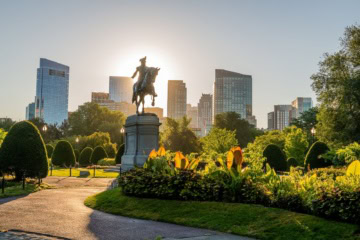 Reiterstatue im Boston Public Garden vor der Skyline der Innenstadt im warmen Abendlicht, umgeben von gepflegten Blumenbeeten und Parkwegen.