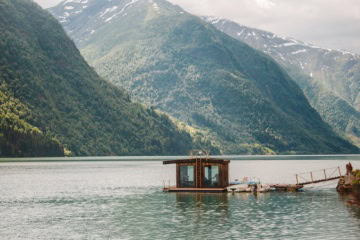 Auf einem See in Norwegen schwimmt eine hölzerne Sauna, mit dem Festland über einen Steg verbunden.