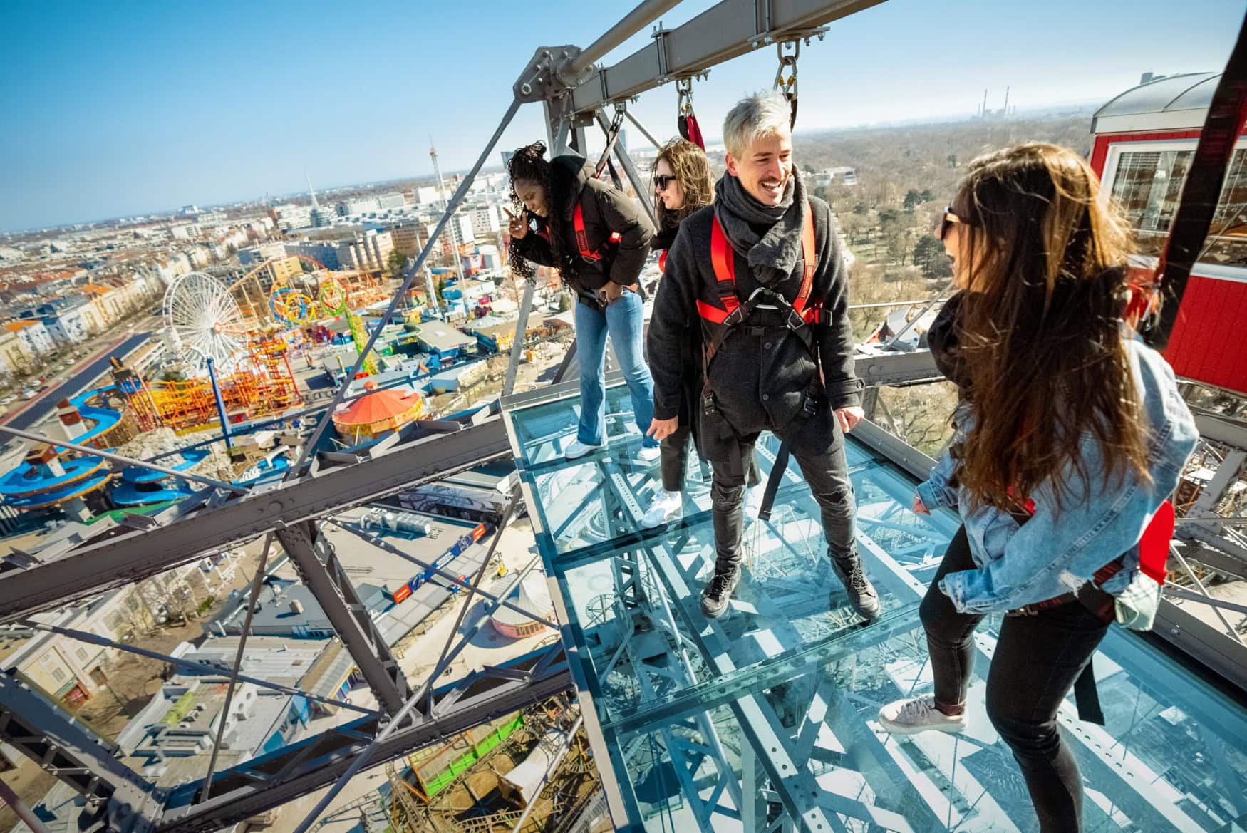 Vier Personen stehen, gesichert mit einem Geschirr, auf einer Plattform, die auf dem Riesenrad am Wiener Prater angebracht ist. Darunter breiten sich die Dächer Wiens aus.
