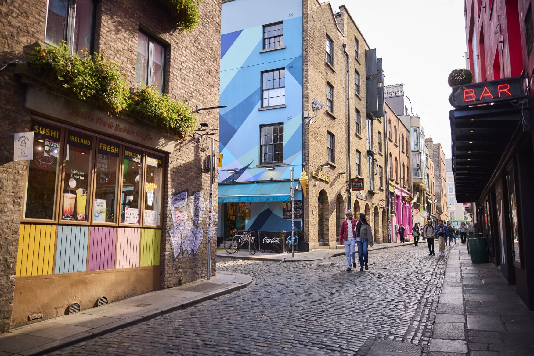 Ein langer, gepflasterter Straßenzug in Dublin. Die Wände der Gebäude sind teils bunt bemalt, auf der linken Seite ist eine Sushibar.
