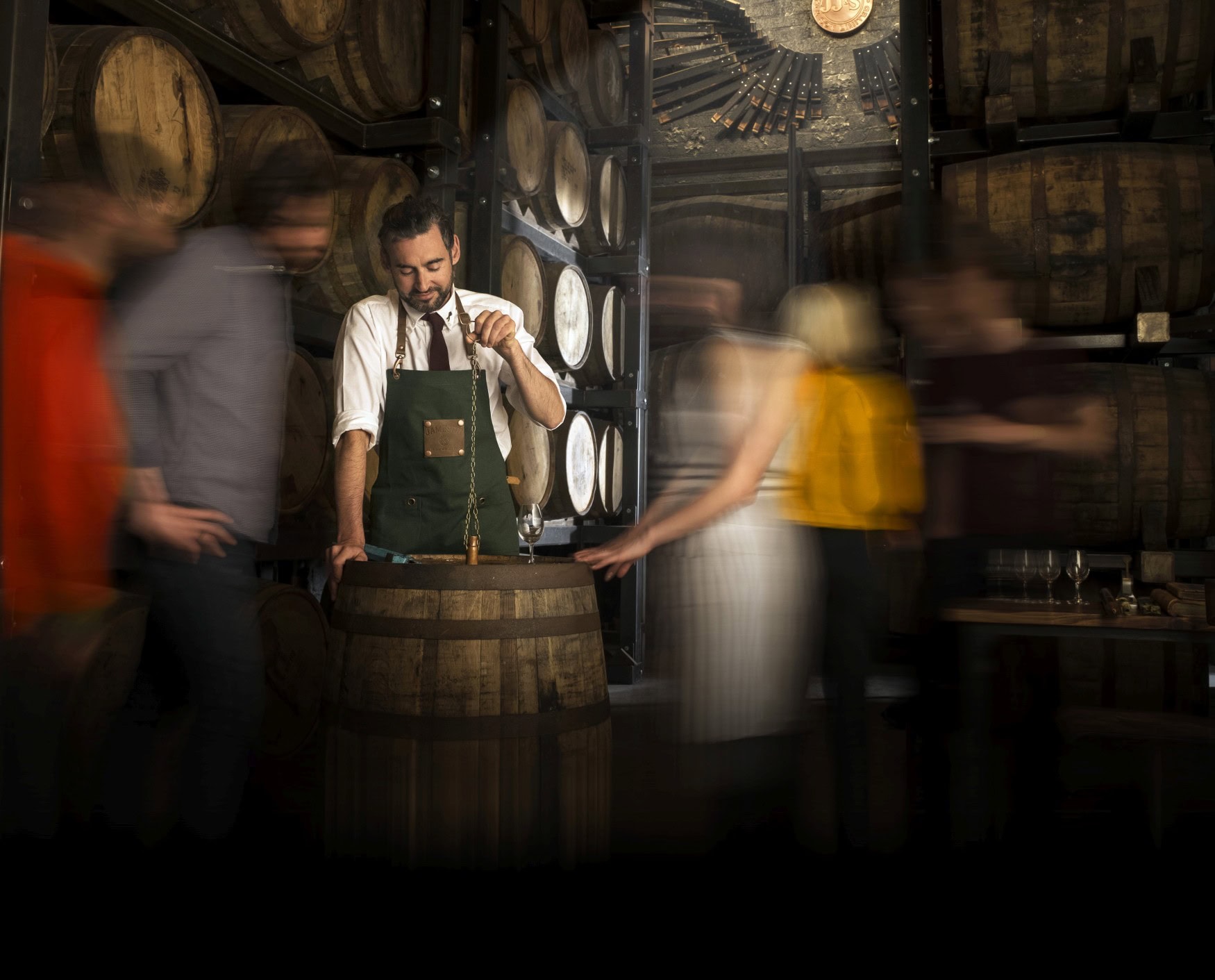 Ein Mann in weißem Hemd und mit grüner Arbeitsschürze steht in einem Lager mit Holzfässern und hebt eine Kette mit kleiner Kupferkelle aus einem hölzernen Fass, auf dem er bereits ein kleines Probierglas bereitgestellt hat. Andere Menschen laufen vorbei, wirken durch Langzeitaufnahme aber verschwommen.