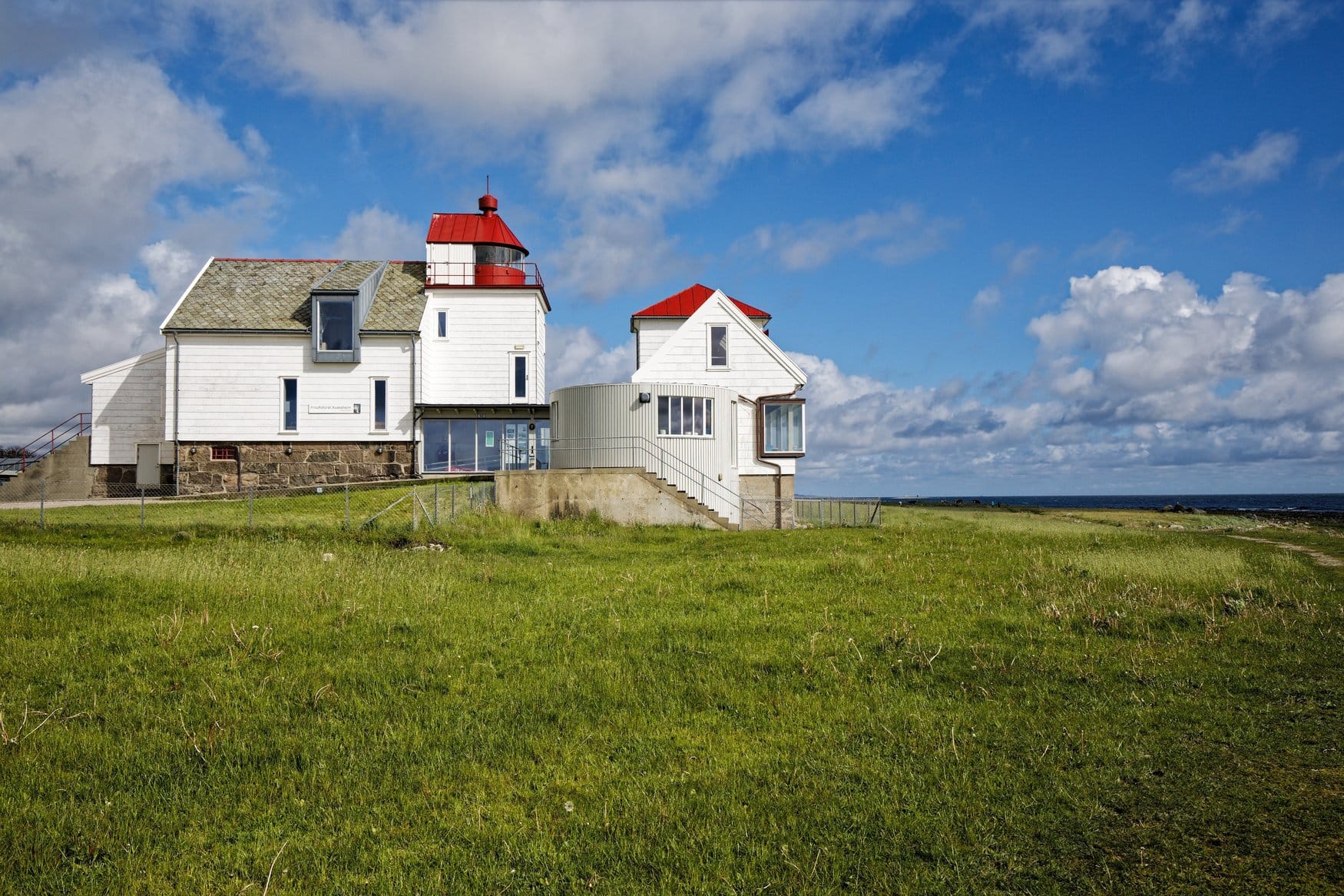 Der weiße Leuchtturm Kvassheim mit rotem Dach steht auf einer grünen Wiese im Jæren in Norwegen, davor ein niedriger angebauter Pavillon mit Rundfenstern.