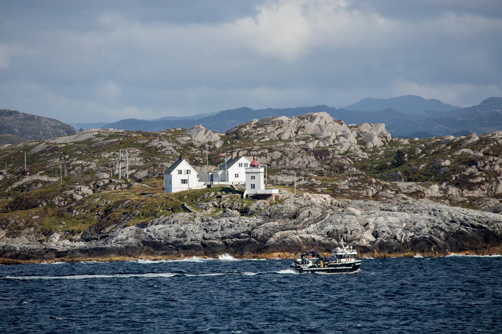 Die weiße Leuchtturmstation Ryvarden auf einer felsigen Landzunge an der norwegischen Küste, davor fährt ein kleines Arbeitsboot vorbei, im Hintergrund Berge unter bewölktem Himmel.