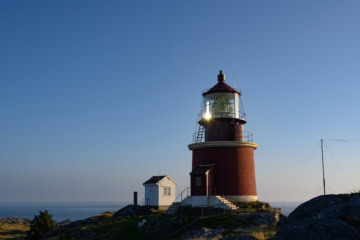 Der weiße Leuchtturm Kvassheim mit rotem Dach steht auf einer grünen Wiese im Jæren in Norwegen, davor ein niedriger angebauter Pavillon mit Rundfenstern.