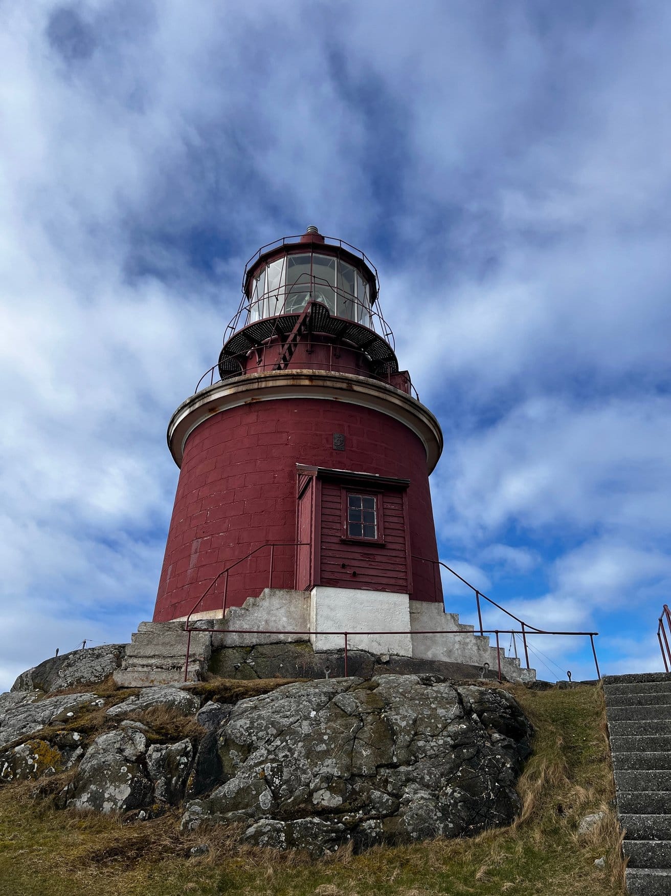 Der dunkelrote Leuchtturm auf Utsira in Norwegen von unten fotografiert, mit Laterne und umlaufendem Balkon, auf einem Felsen stehend unter bewölktem Himmel.