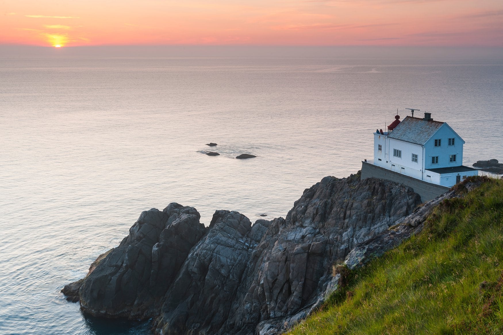 Der Leuchtturm Kråkenes auf einer Klippe bei Vågsøy in Norwegen, im Vordergrund schroffe Felsen und ruhige See, am Horizont geht die Sonne unter.
