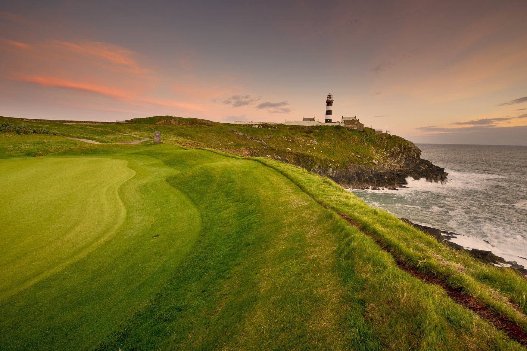 Der schwarz-weiß gestreifte Leuchtturm am Old Head of Kinsale in der Grafschaft Cork bei Abendlicht. Im Vordergrund liegt der Rasen eines Golfplatzes auf den Klippen, rechts brandet das Meer. 