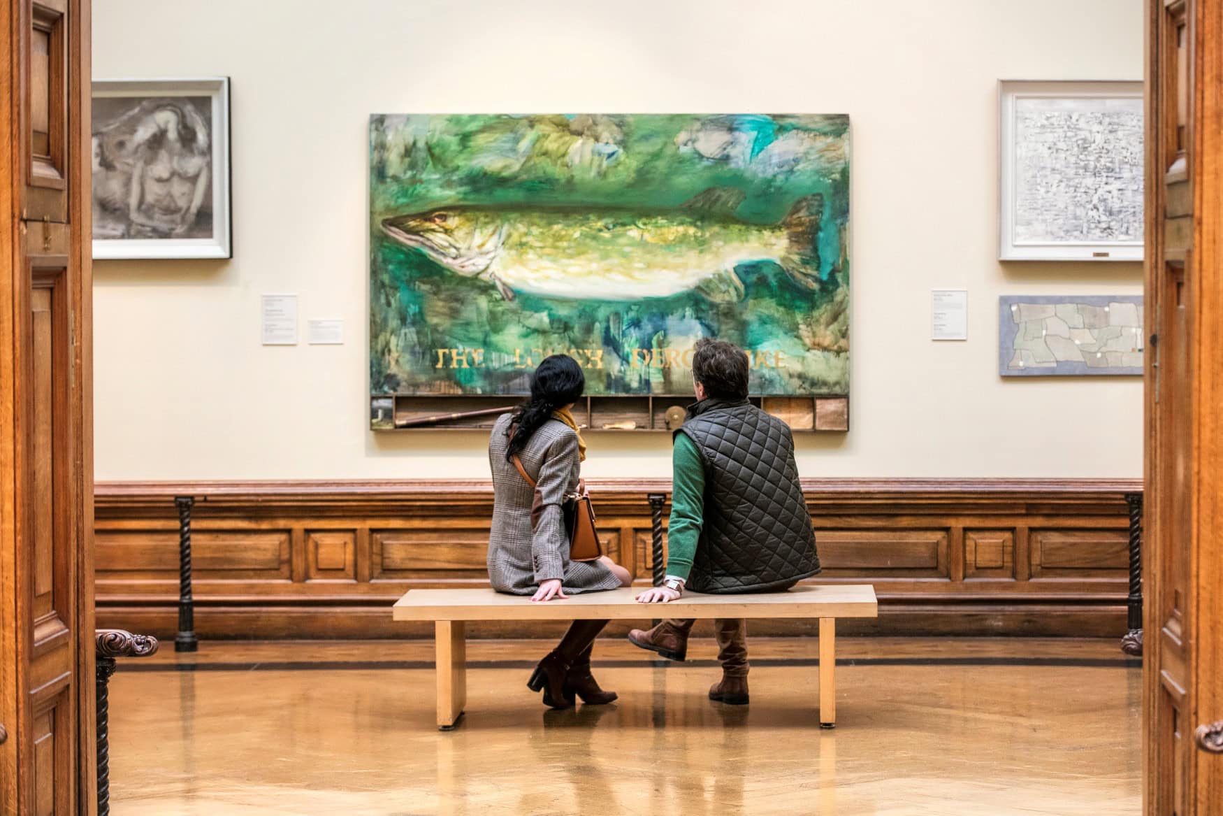 Zwei Menschen sitzen auf einer Bank in einer Galerie und betrachten einen modernen Kunstdruck, der einen Hecht zeigt.