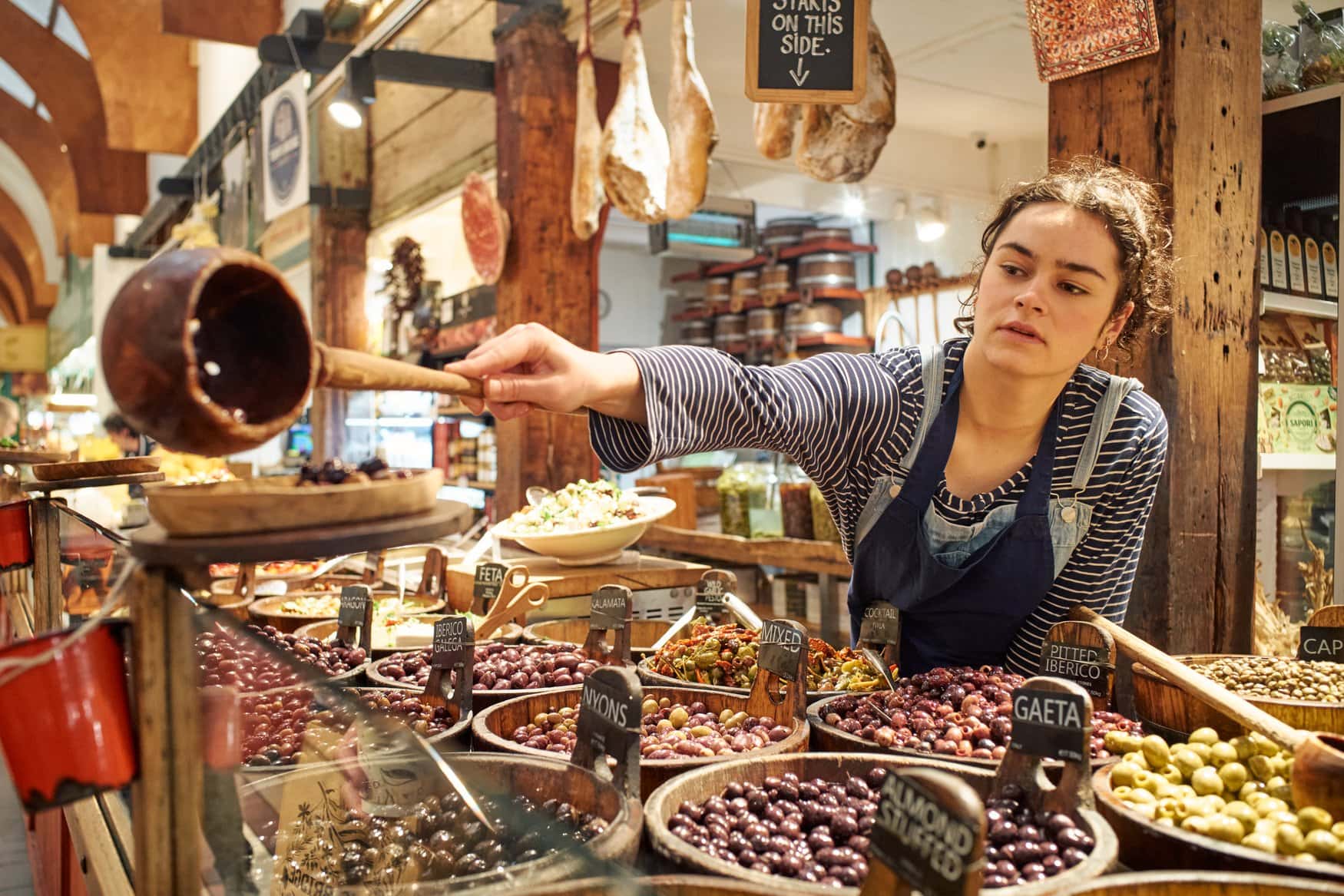 Eine Verkäuferin schöpft an einem Stand im English Market in Cork Oliven aus einer Holzschale. Vor ihr stehen zahlreiche Schalen mit verschiedenen Oliven, darüber hängen luftgetrocknete Schinken.
