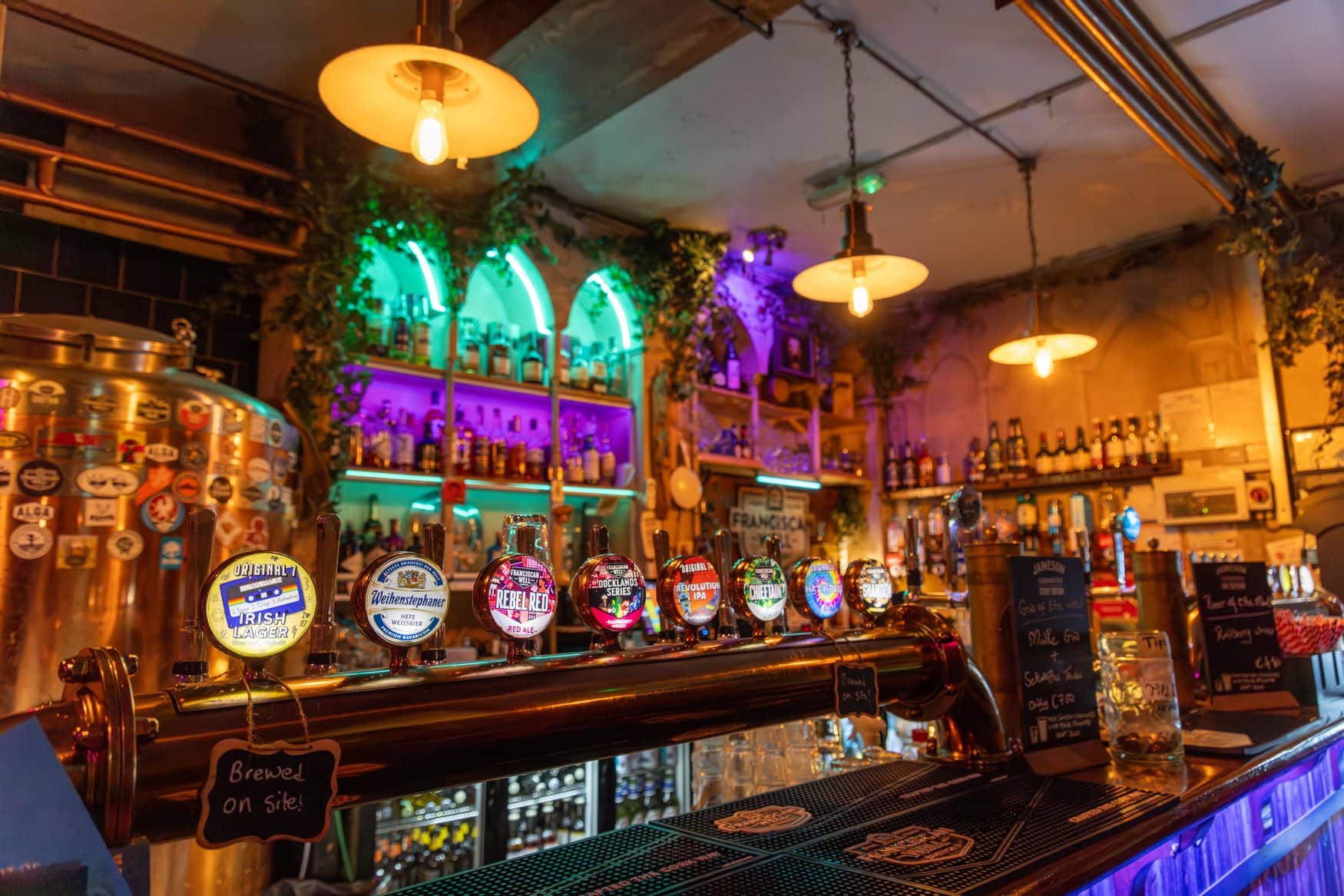 Zapfanlage im Brewpub der Franciscan Well Brewery in Cork. Über der Theke mit mehreren Bierhähnen hängen Lampen, dahinter stehen ein kupferner Braukessel und bunt beleuchtete Flaschenregale. 