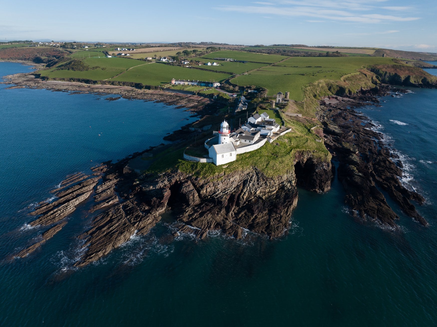 Der rot-weiße Leuchtturm am Roches Point am Eingang zum Hafen von Cork. Er steht mit einigen weißen Gebäuden auf einer felsigen Landzunge, umgeben von Feldern und Meer.
