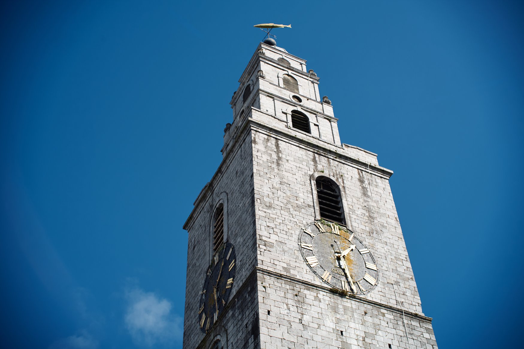 Der Turm der St Anne's Church im Stadtteil Shandon in Cork vor blauem Himmel. Auf der Spitze sitzt eine goldene Fischfigur als Wetterfahne, darunter sind die Zifferblätter der Turmuhr zu sehen.