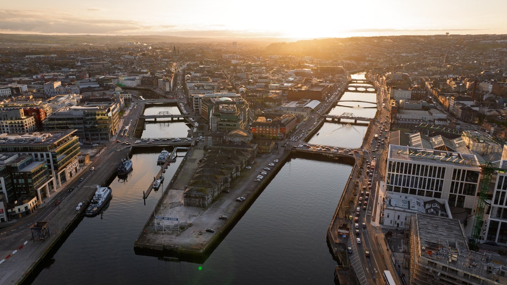 Luftaufnahme der Innenstadt von Cork bei Sonnenuntergang. Der Fluss Lee teilt sich in zwei Arme, die von mehreren Brücken überspannt werden, am Kai liegt der Port of Cork. 