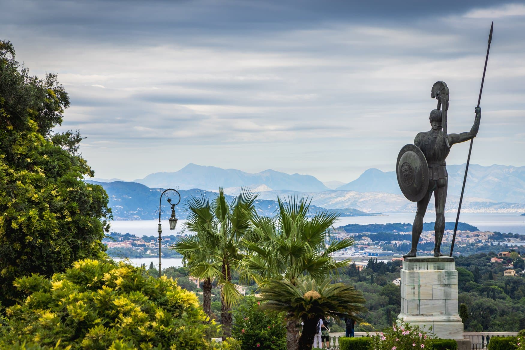 Die Bronzestatue des Achilles im Garten des Achilleion auf Korfu, von hinten aufgenommen. Über Palmen hinweg fällt der Blick auf die Bucht und die Berge des Festlands.