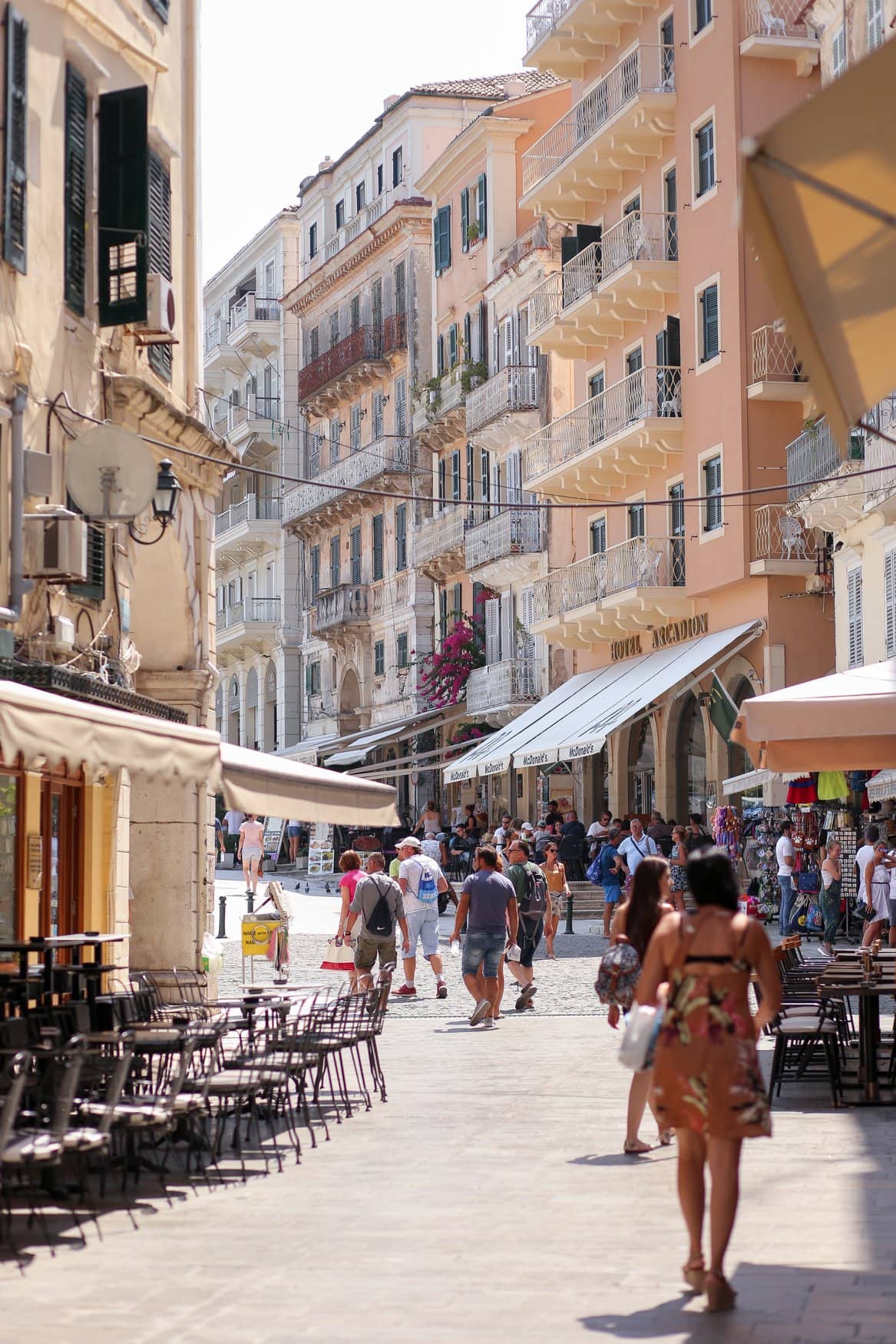 Eine Gasse in der Altstadt von Korfu mit hohen Häusern und schmiedeeisernen Balkonen. Passanten gehen zwischen Ladenzeilen und Tischen der Straßencafés hindurch.