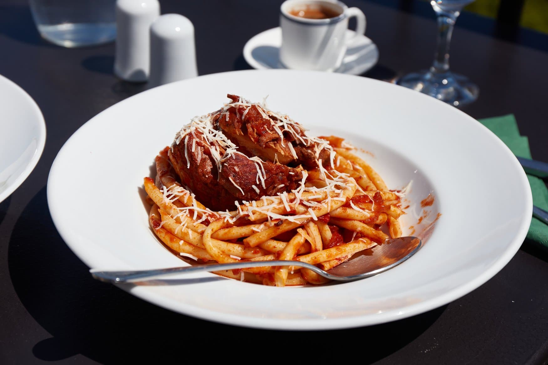 Ein Teller Pastitsada: geschmortes Rindfleisch auf Bucatini in Tomatensauce, bestreut mit geriebenem Hartkäse. Der Teller steht auf einem dunklen Tisch im Freien.