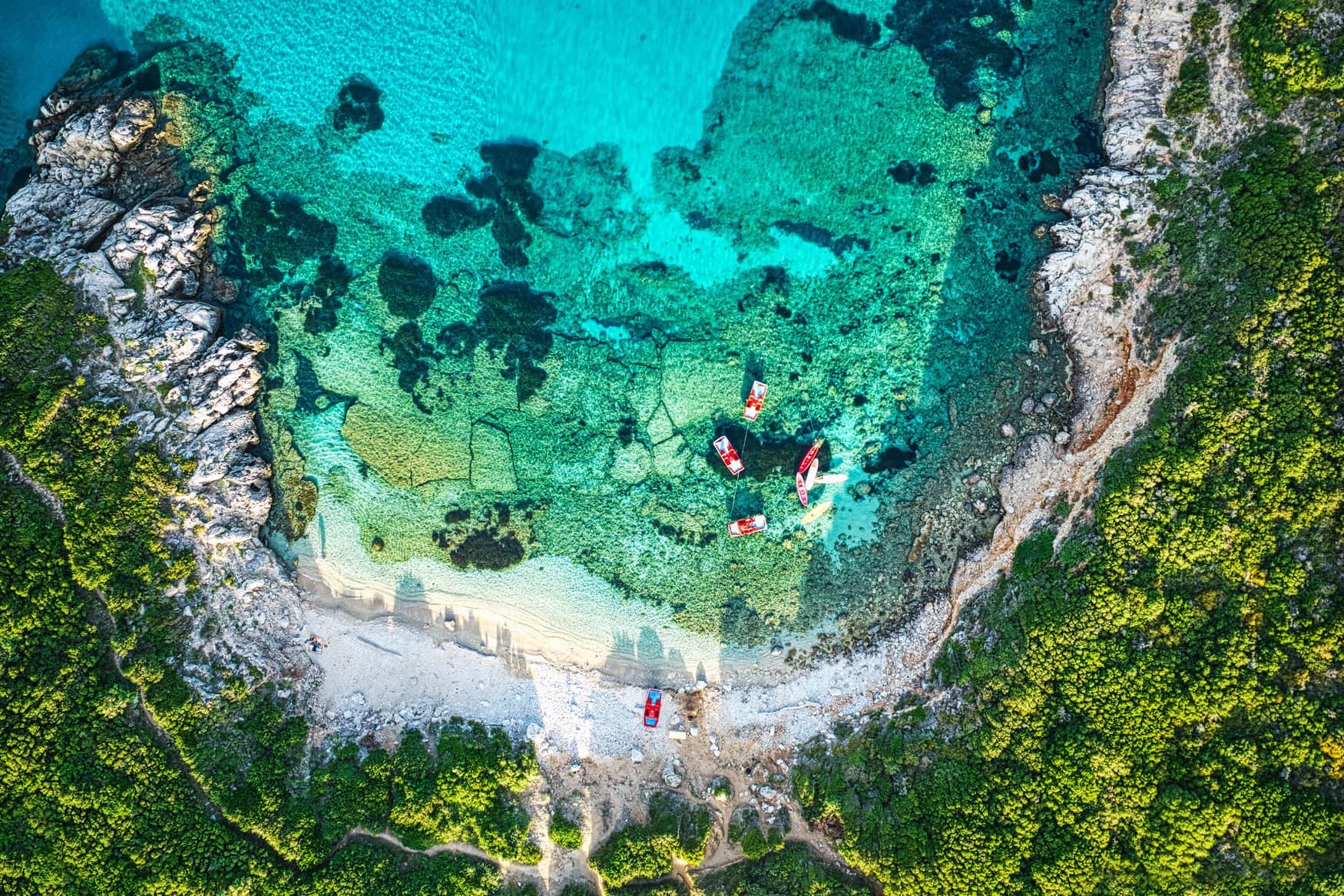 Senkrechte Luftaufnahme der Bucht Porto Timoni auf Korfu mit türkisfarbenem Wasser über hellem Grund. Am Kiesstrand liegen Boote, ringsum steigen bewachsene Felshänge an.