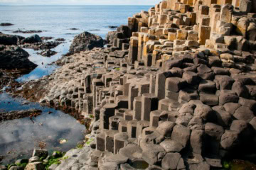 Sechseckige Basaltsäulen bilden eine zerklüftete Steinküste an der nordirischen Atlantikküste, im Vordergrund liegt grünes Seegras zwischen den Felsen.