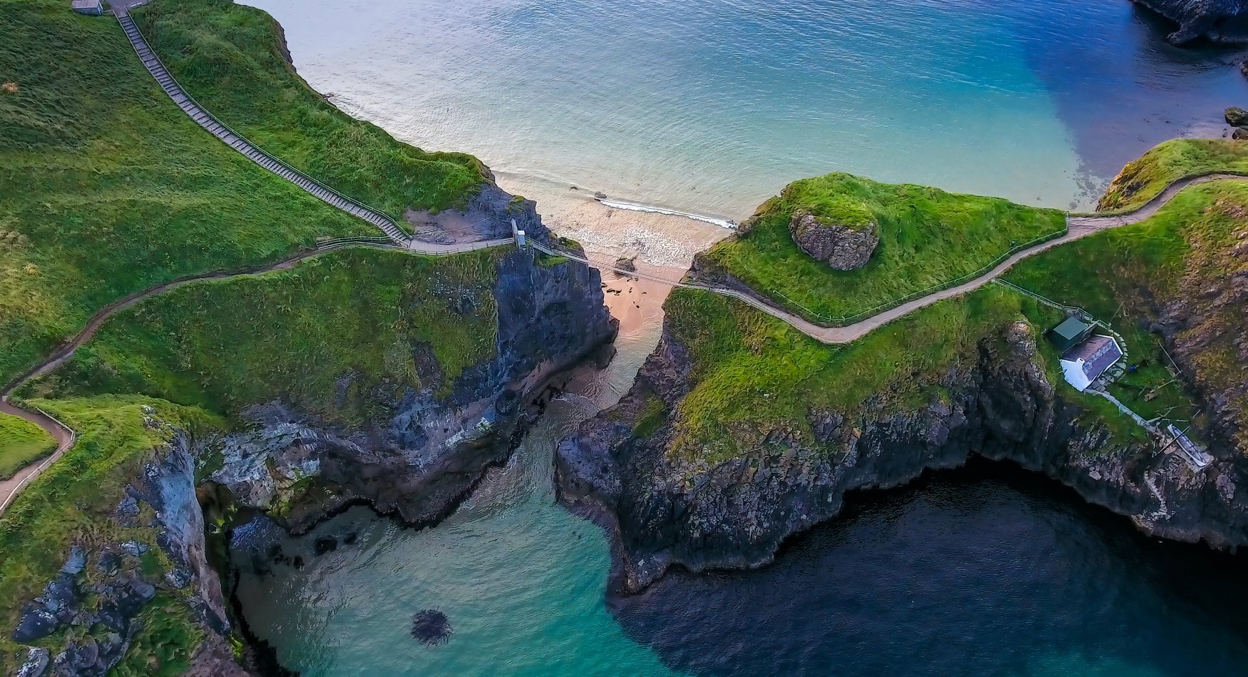 Luftaufnahme einer grünen Landzunge bei Carrick-a-Rede in Nordirland mit einem schmalen Sandstrand zwischen zwei Klippen und türkisfarbenem Wasser.