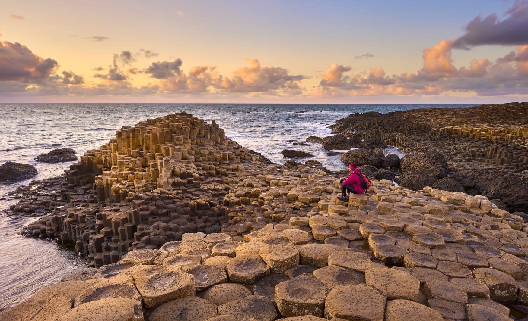 Eine Person sitzt bei Sonnenuntergang auf den sechseckigen Basaltsäulen des Giant's Causeway in Nordirland und blickt aufs Meer, am Horizont ziehen rosa gefärbte Wolken.