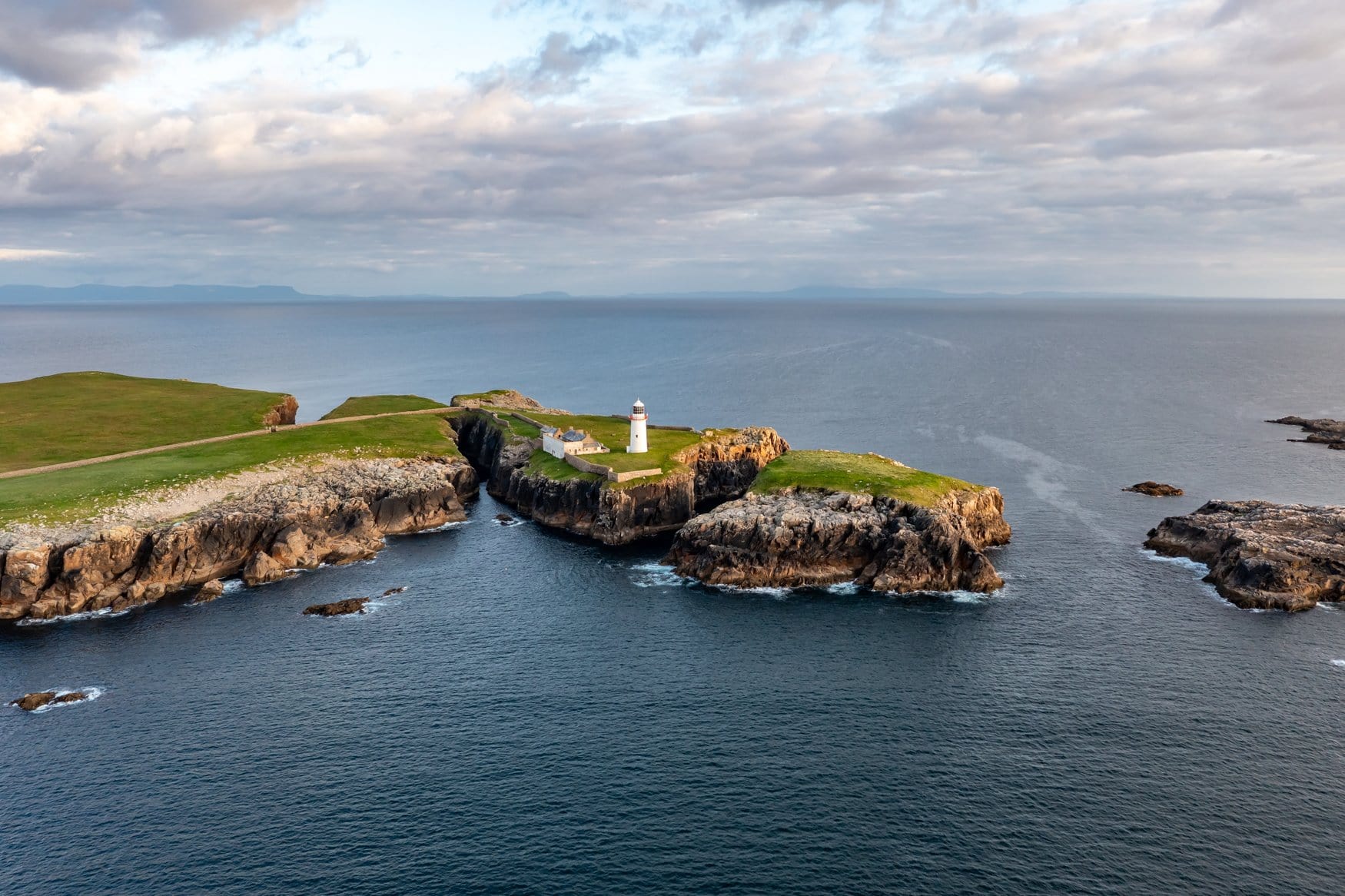 Luftaufnahme eines weißen Leuchtturms auf einer schmalen Landzunge bei Rathlin Island in Nordirland, die Klippen fallen steil zum Meer ab, im Hintergrund die Küste des Festlands.