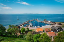 Ausblick von einem grünen Hügel auf einen kleinen Yacht-Hafen auf einer Ostseeinsel. Der Himmel über der Szene ist sommerlich-blau.