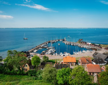 Ausblick von einem grünen Hügel auf einen kleinen Yacht-Hafen auf einer Ostseeinsel. Der Himmel über der Szene ist sommerlich-blau.