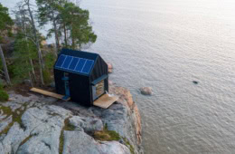 Am felsigen Ufer eines Sees steht eine kleine Lodge aus schwarzem Holz, auf dem Dach ein Solar-Panel.