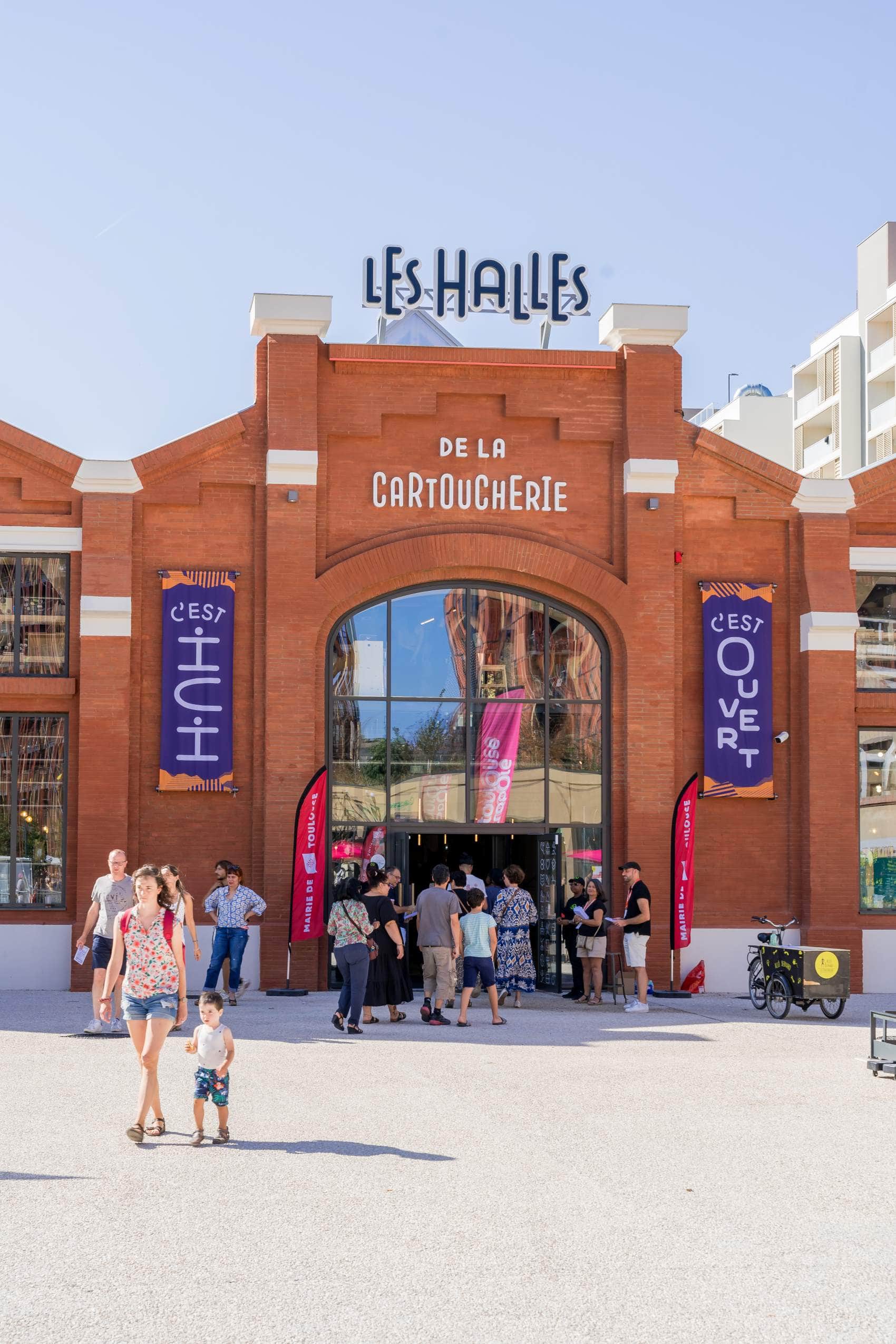 Les Halles de la Cartoucherie in Toulouse, ein umfunktioniertes Fabrikgebäude aus Backstein, davor eine Schlange an Menschen in sommerlicher Kleidung.
