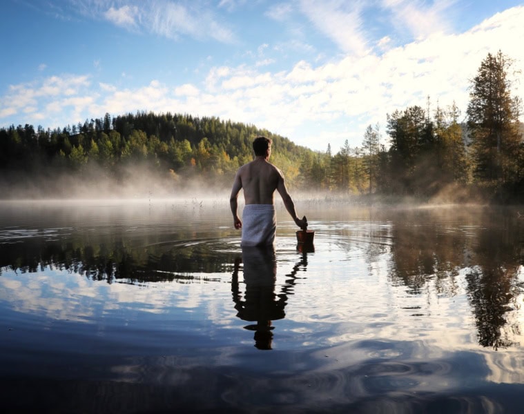 Ein Mann steht, nur mit einem Handtuch bekleidet und einem Holzeimer in der Hand, bis zu den Oberschenkeln in einem weiten See. Auf der gegenüberliegenden Uferseite beginnt ein dichter Nadelwald.
