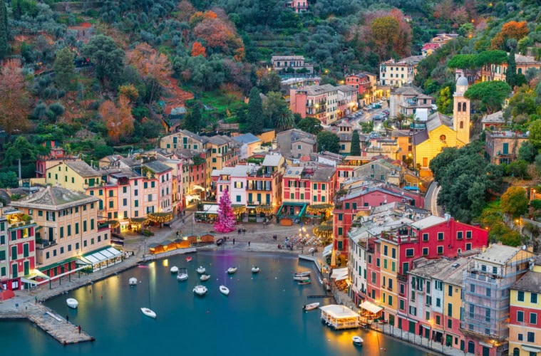 Luftaufnahme des Hafens von Portofino in der Abenddämmerung mit bunten Pastellhäusern, beleuchtetem Weihnachtsbaum auf der Piazzetta und ankernden Booten.
